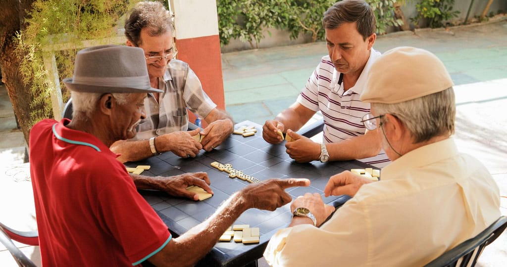 Friends Sharing a Game of Dominoes Outdoors - The Preserve Four older adults sitting together at a table outside, smiling and connecting over a game of dominoes.