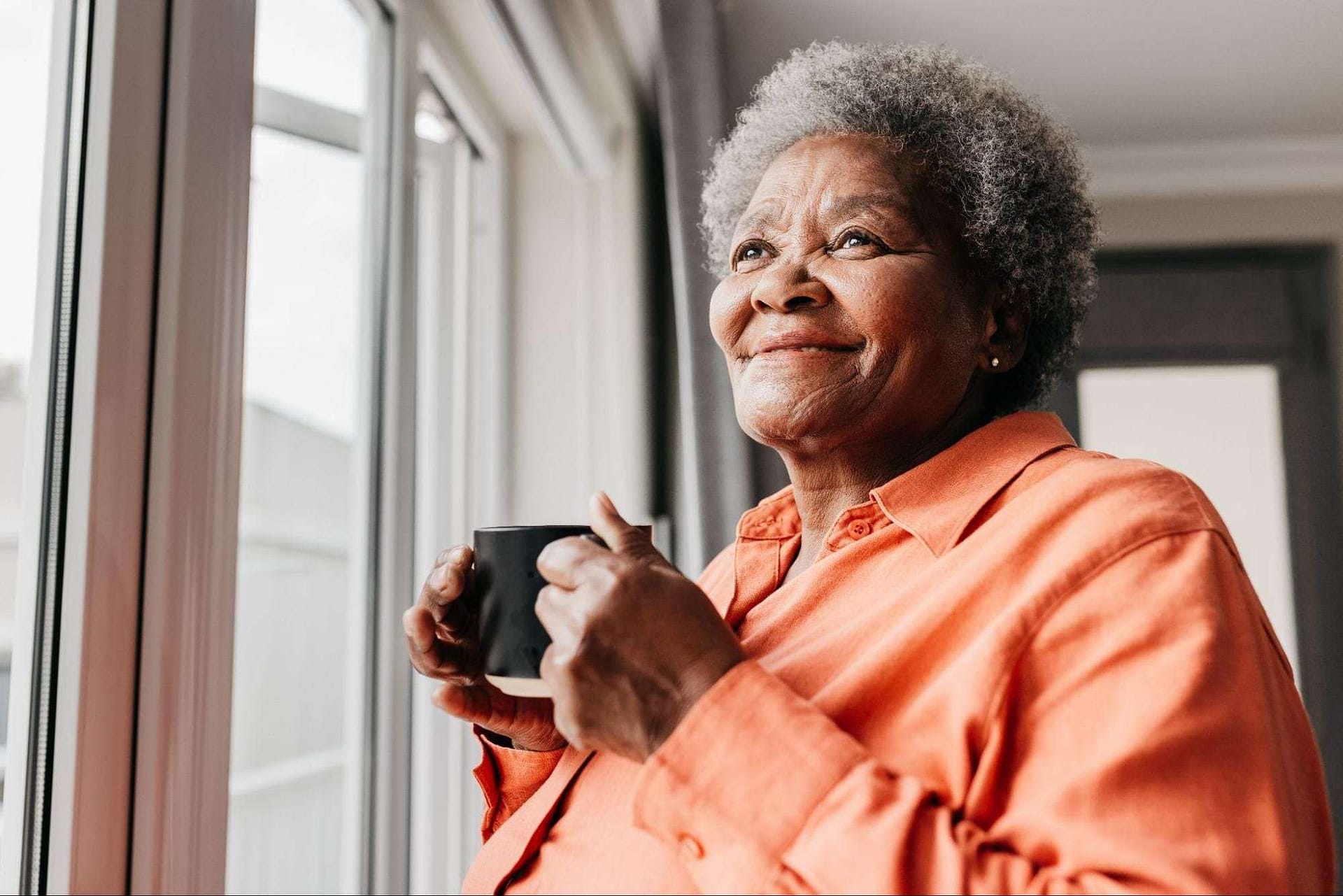 Woman Enjoying Morning Coffee - The Preserve A woman smiles while holding a coffee mug, looking out a bright window, while thinking about senior living options.